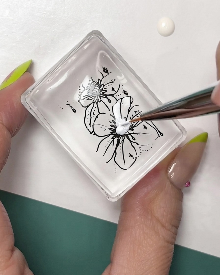 Ein feiner Liner-Pinsel fügt weiße Details zu schwarz umrandeten Blumenstempeln auf einem klaren Stempelkissen hinzu.