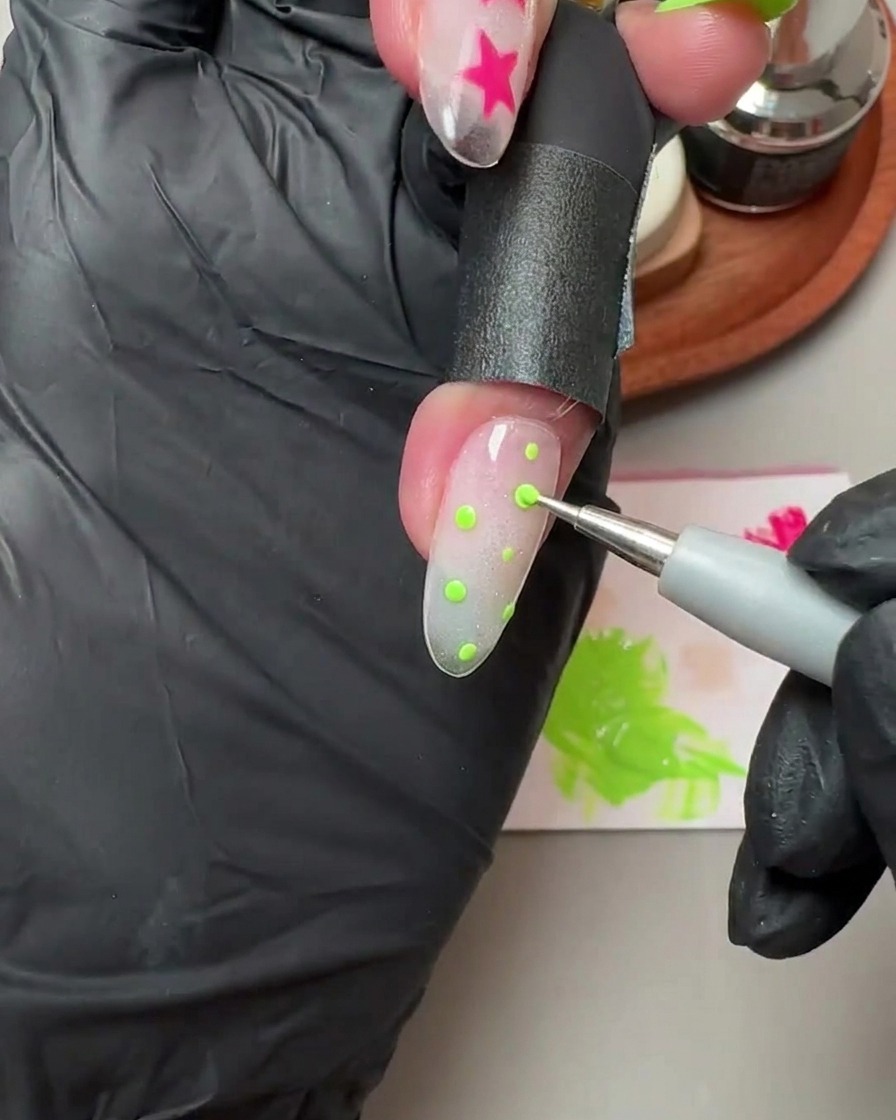 Ein Dotting-Tool platziert kleine grüne Polka Dots auf einem durchscheinenden Glitter- und Cat-Eye-Gel-Nagel.