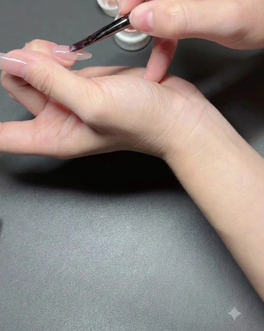 Ein Nagelpinsel trägt eine dünne Schicht Nude semi-sheer Nagellack auf einen Fingernagel auf.