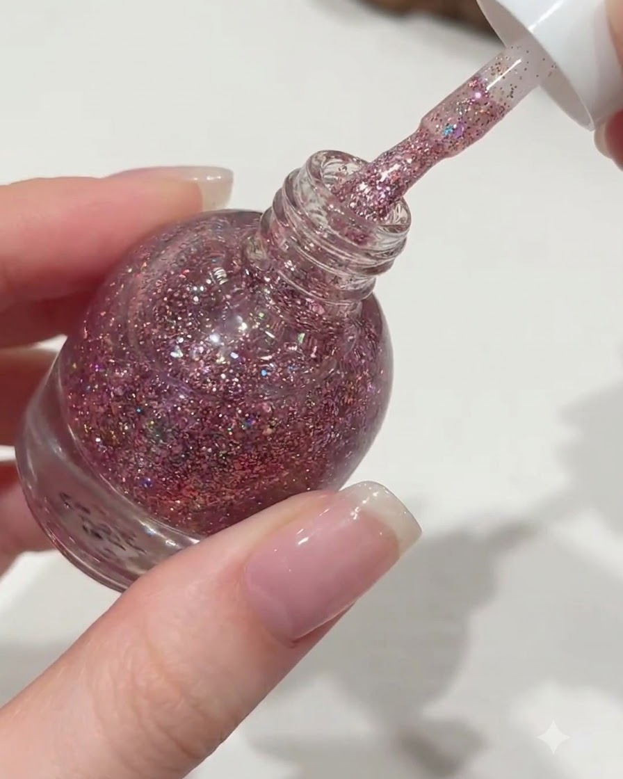 Eine Hand schraubt den weißen Deckel einer Orange Nagellackflasche auf und zieht einen Pinsel heraus, der mit pinkem Glittergel benetzt ist.