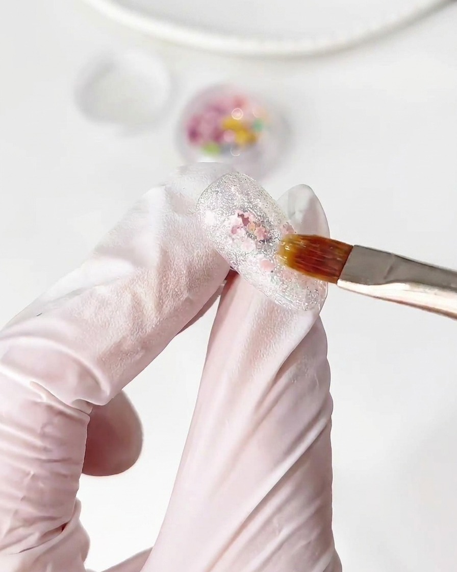 Ein Pinsel platziert kleine, bunte Blumen-Glitzerteile auf einer nassen Base-Gel-Schicht auf einem Nagel.