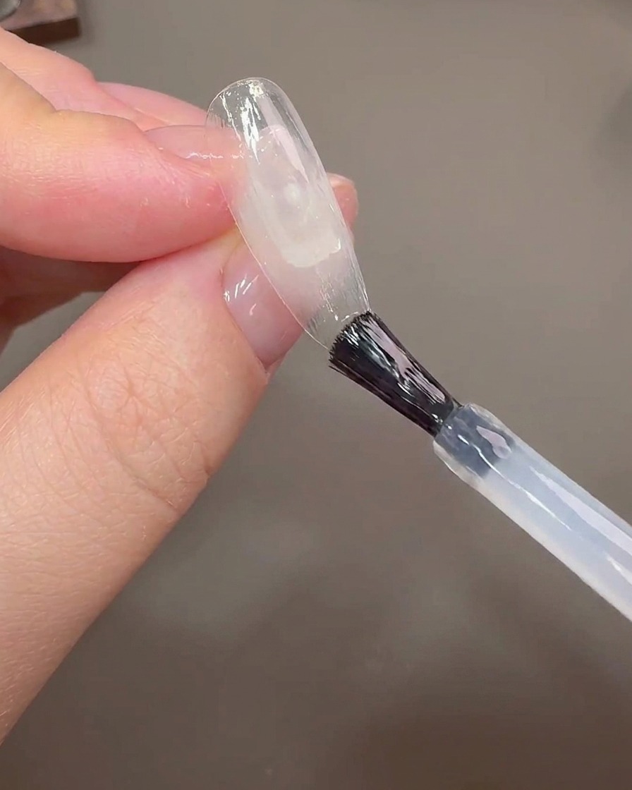 Ein Nagelpinsel trägt Aufbaugel auf eine klare Nagelspitze auf, um die Wachstumslinie abzudecken.