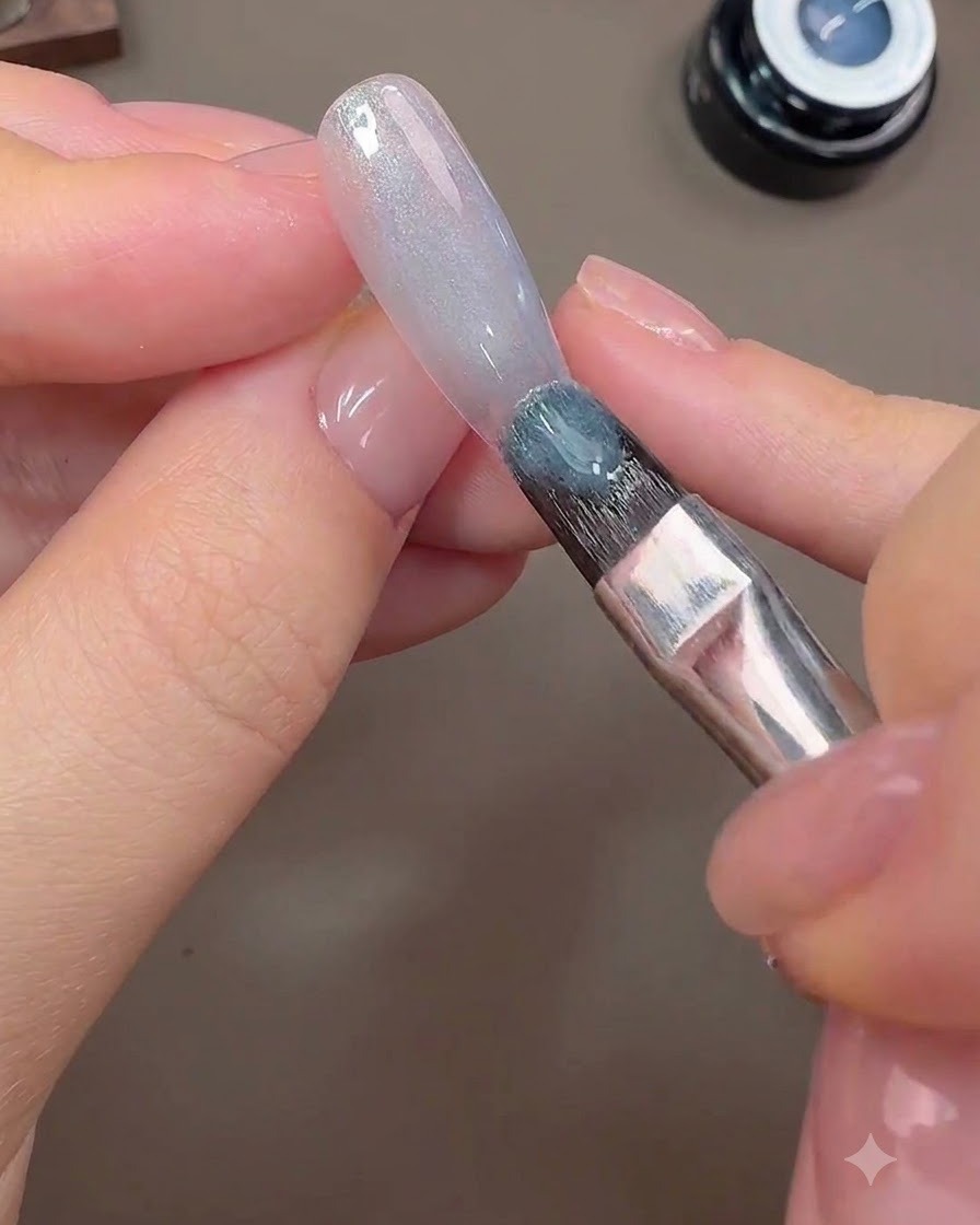 Ein Pinsel trägt grau-blautes Cat-Eye-Gel auf die Nageloberfläche auf, um den galaktischen Hintergrund zu bilden.