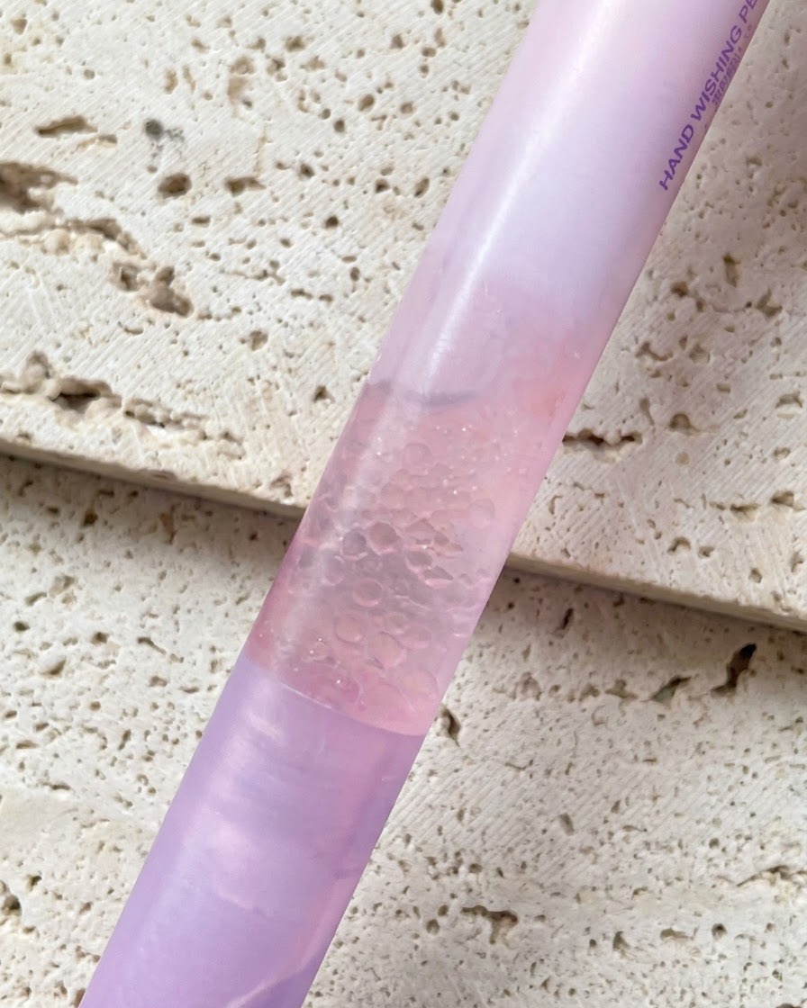 Ein rosa Perlmutt-Gradienten-Nagelölstift wird vor einem strukturierten Hintergrund gehalten.
