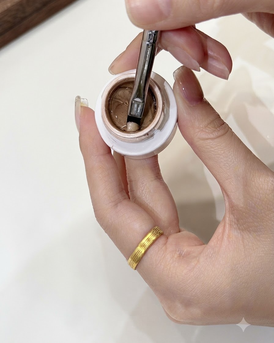 Ein Nail-Art-Pinsel nimmt eine kleine, bohnengroße Portion Champagner-Gold-Gel aus einem offenen Tiegel auf.