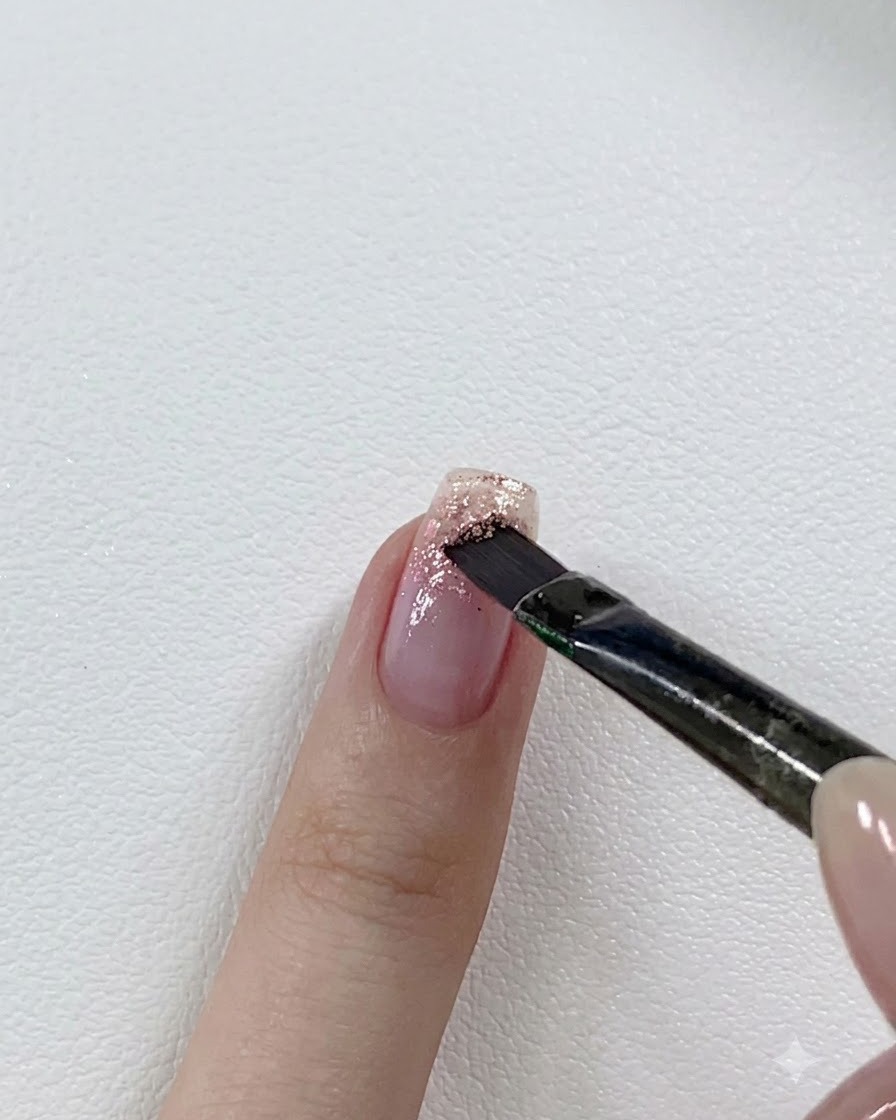 Ein Nagelpinsel schichtet einen dunkleren rosa Glitzer-Gel-Ton über die Spitze eines teilweise glitzernden Nagels.