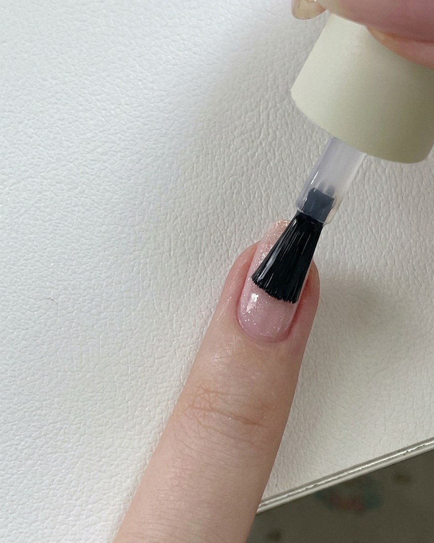 Ein Pinsel trägt einen klaren, glänzenden Top Coat über einen rosa Glitzer-Gel-Nagel auf.