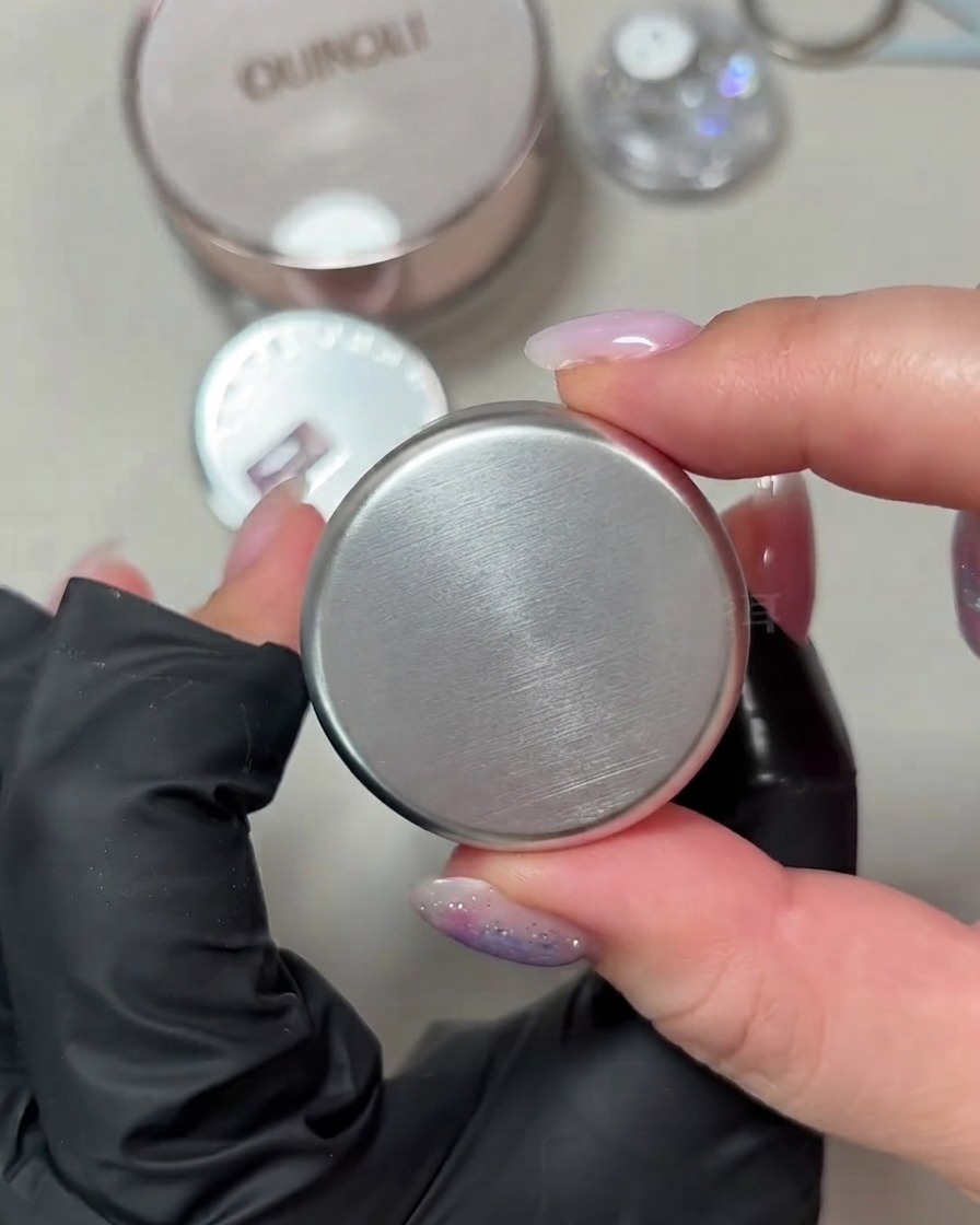 Eine Hand hält einen kleinen runden Tiegel mit porzellanweiß schimmerndem Cat-Eye-Gel vor einem neutralen Hintergrund.