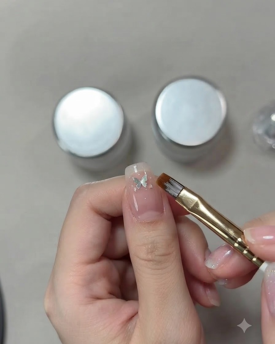 Eine detaillierte Nahaufnahme der Hand einer Nail Artistin, die mit einem feinen Pinsel eine kleine silberne Schmetterlings-Paillette auf einem rosa Cat-Eye-Nagel positioniert.