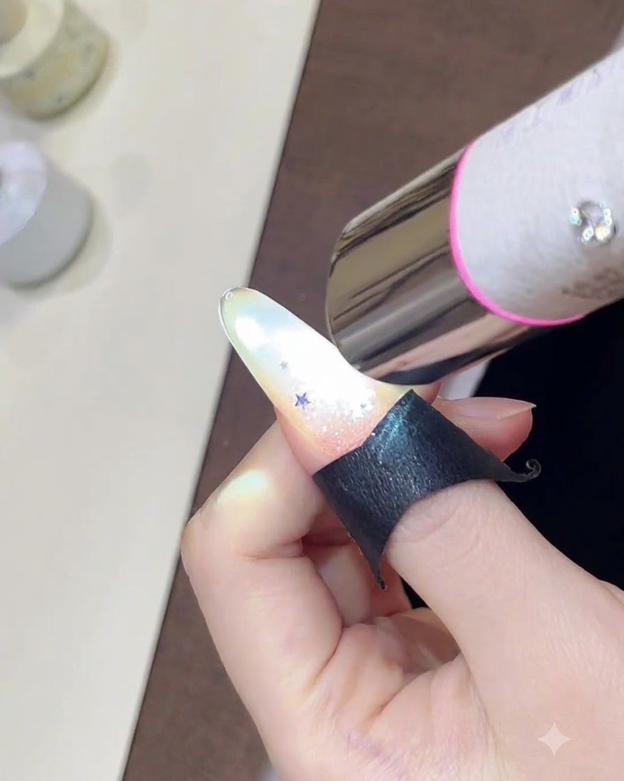 Eine Hand hält eine Mini-UV-Lampe über einen Nagel, der mit einem funkelnden Glitzer-Farbverlauf und kleinen Stern-Pailletten verziert ist.