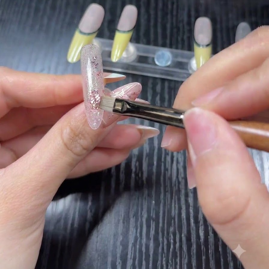 Nahaufnahme eines Pinsels, der rosa und silberne Hologramm-Glitterflocken über einer transparenten Basis auf eine Übungs-Nagelspitze aufträgt.