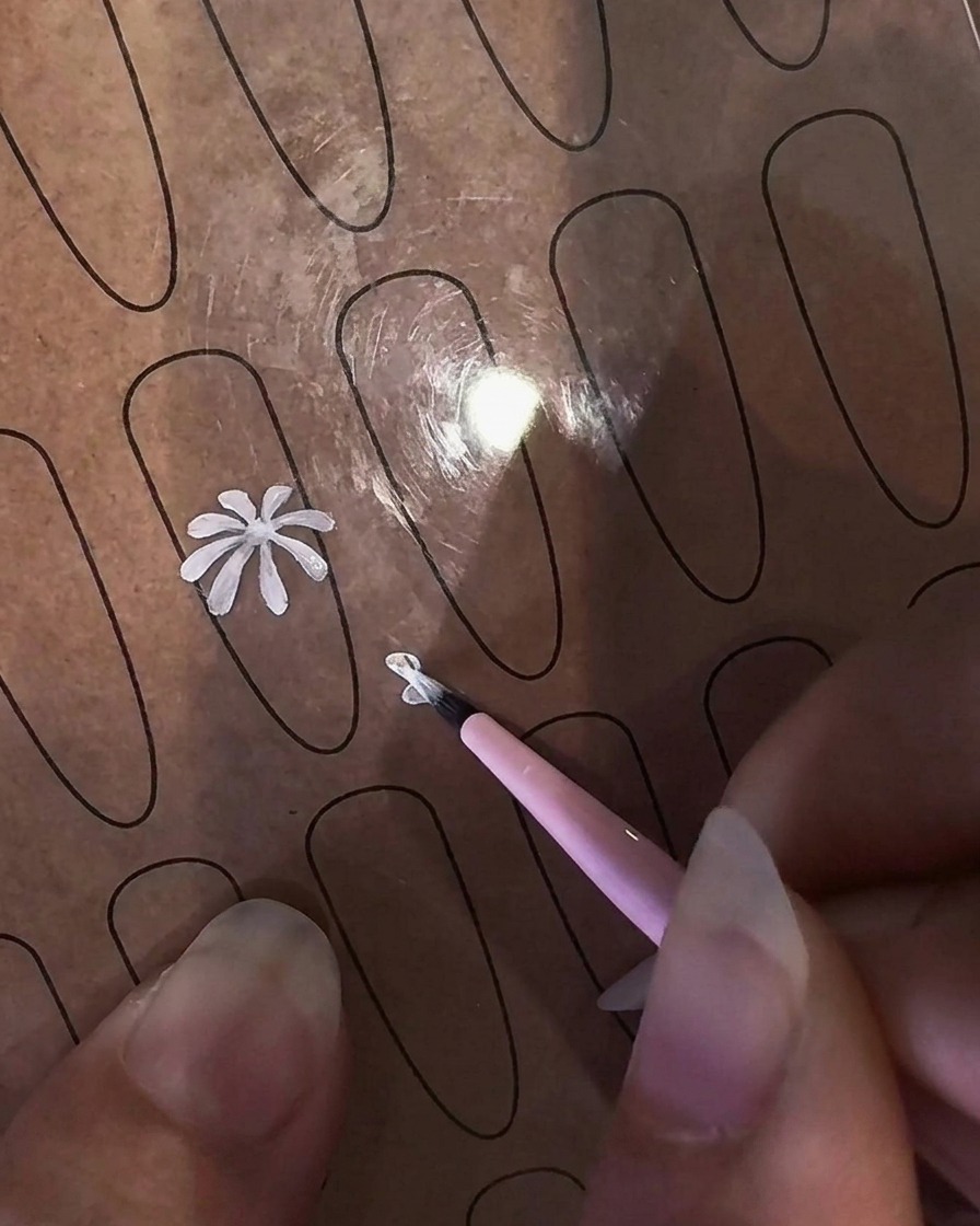 Nahaufnahme einer Hand, die einen feinen Liner-Pinsel hält, um ein weißes Gänseblümchen-Blütenblatt auf ein klares Nagel-Übungsblatt zu malen.