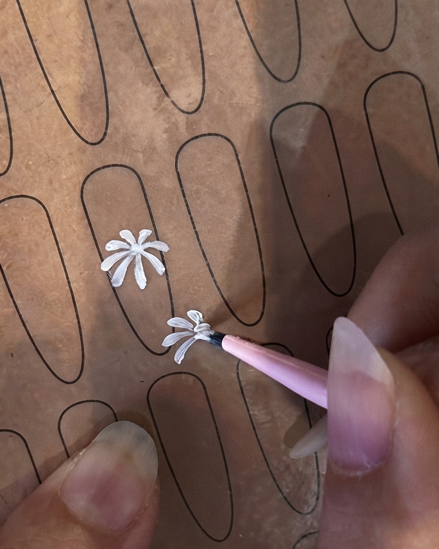 Ein feiner Liner-Pinsel beginnt mit einem zweiten, kleineren weißen Blütenblatt auf einem Nagel-Übungsblatt.