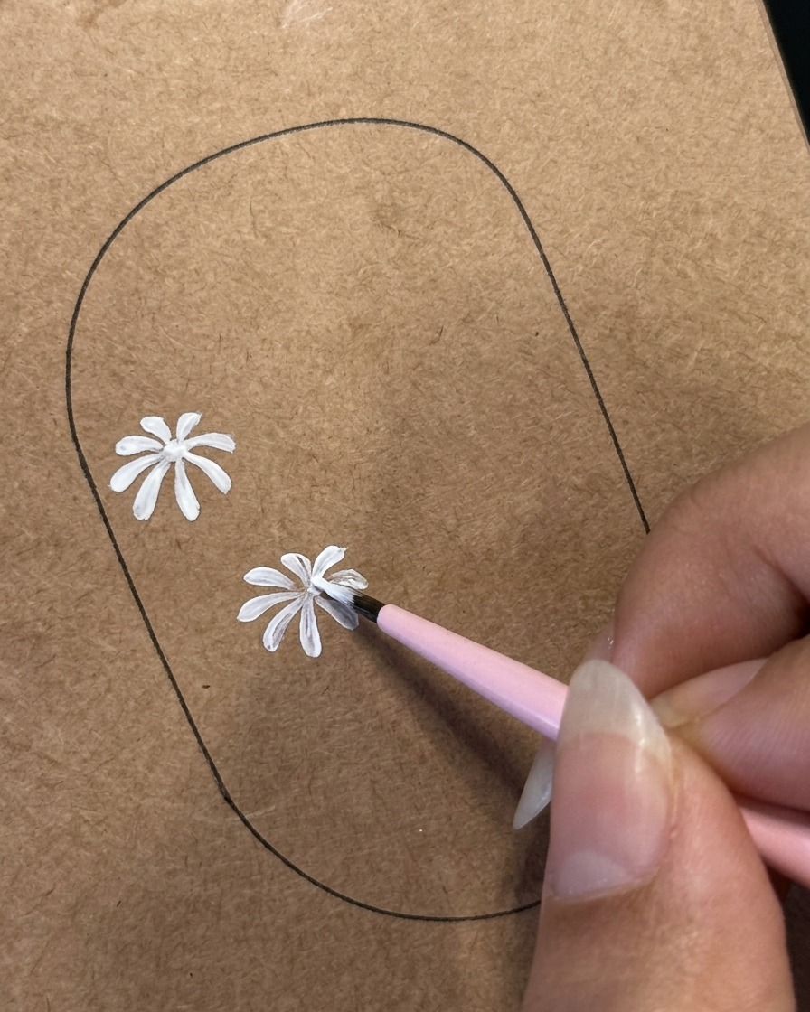 Ein feiner Liner-Pinsel detailliert weiße Blütenblätter an einem zweiten Gänseblümchen-Design, das auf eine braune Nagel-Übungsschablone gemalt wurde.