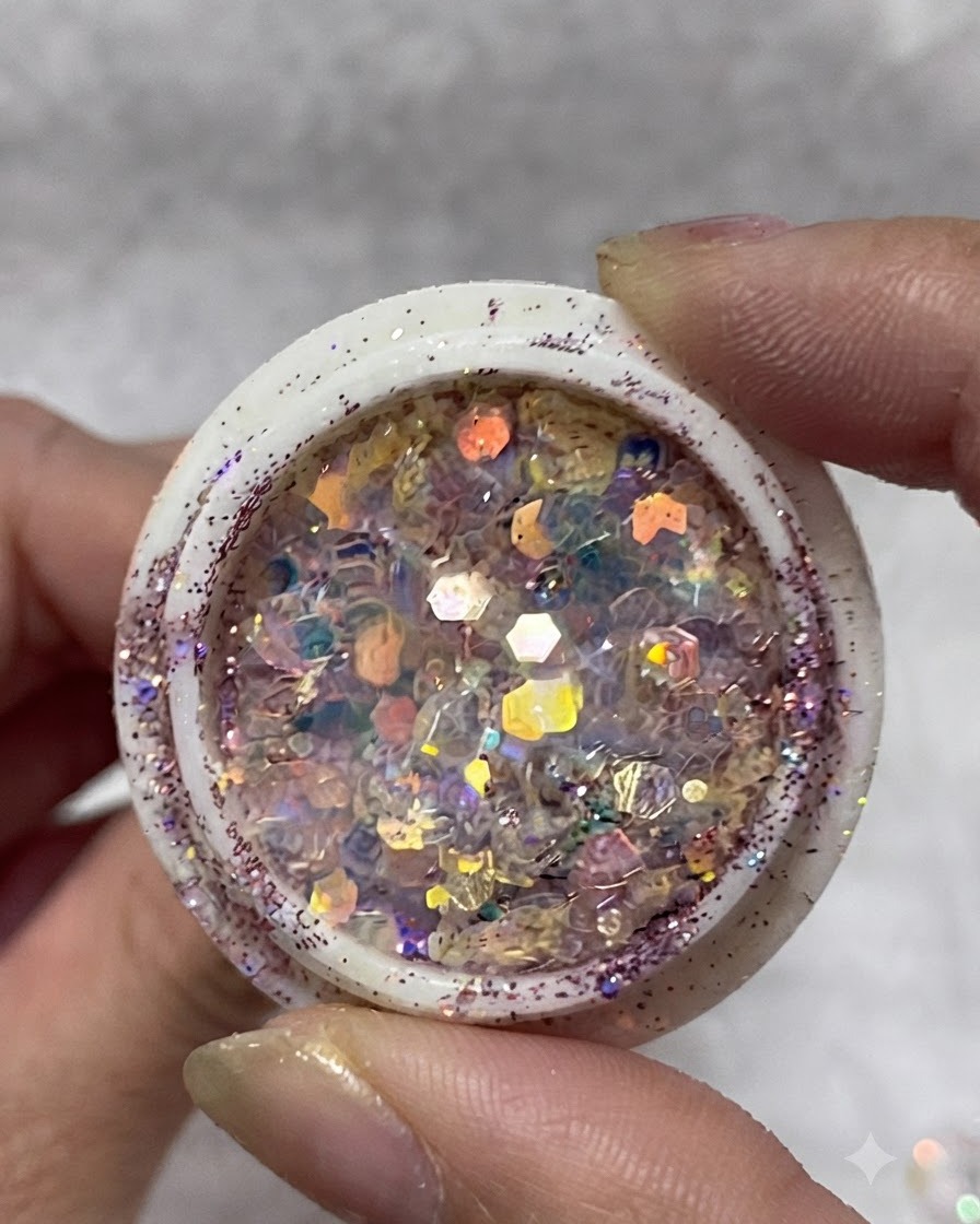 Ein Tiegel mit mehrfarbigen, irisierenden Glitzerflocken, Pailletten und kleinen Sternen, gehalten zwischen den Fingern.