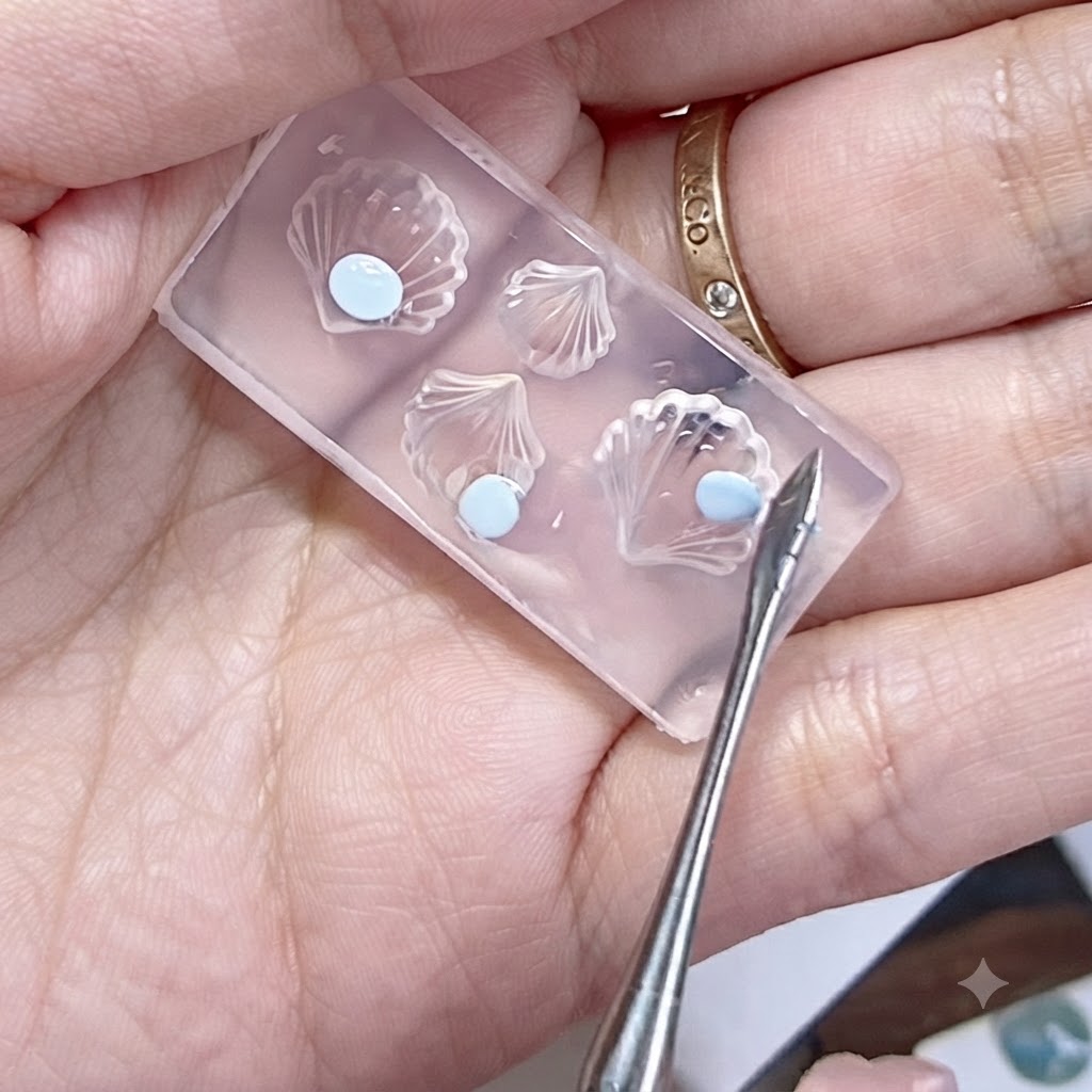 Eine Silikonform in einer Hand mit flachen blauen Gelperlen und einem Werkzeug daneben.