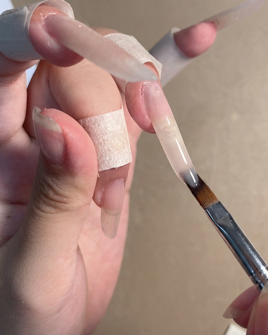 Ein Nagelpinsel trägt ein transparentes Nude-Gel-Polish auf eine lange Stiletto-Nagelverlängerung auf.