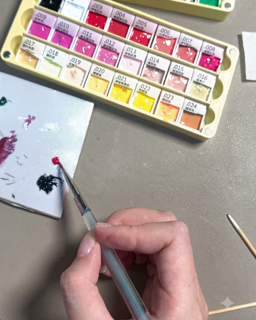 Eine Hand verwendet einen feinen Detailpinsel, um einen kleinen Tropfen helles Rosenrot-Gel auf eine weiße Keramik-Mischpalette aufzutragen.
