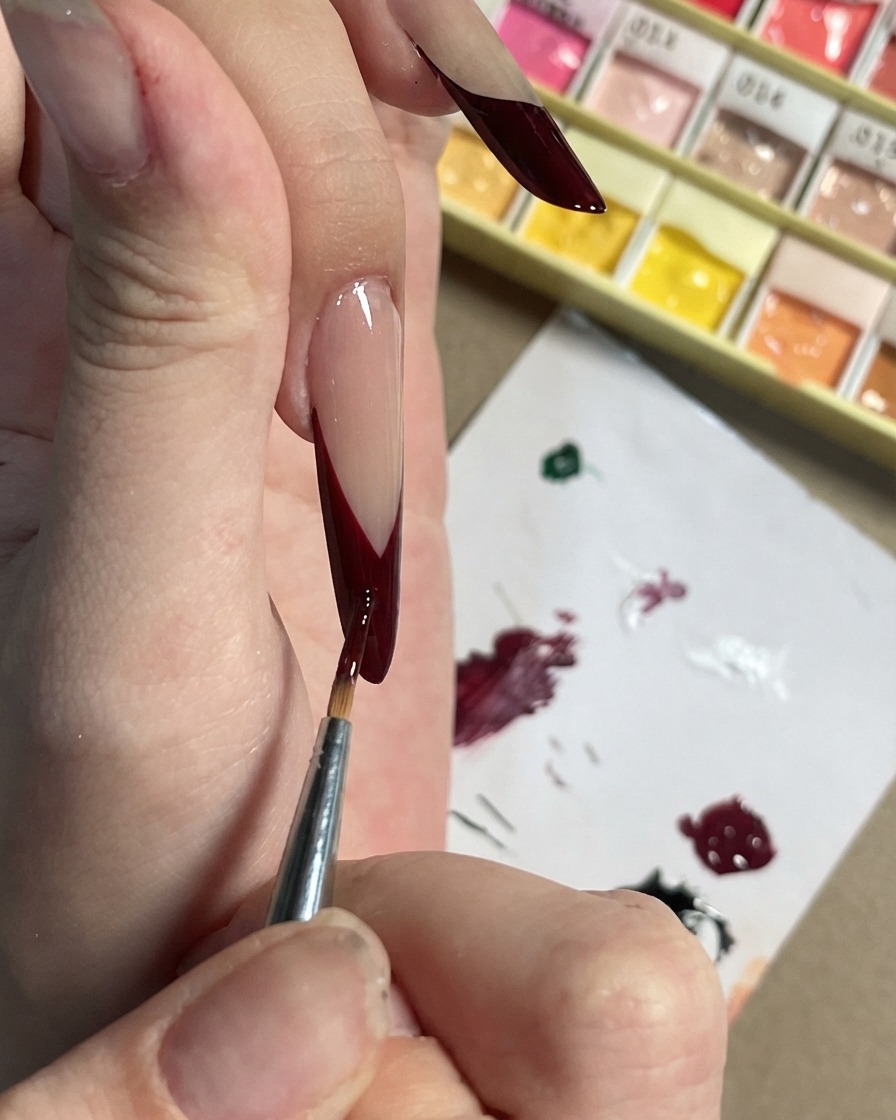 Ein feiner Pinsel fügt kleine dunkelviolette Akzente und Blütenblattformen auf einer schwarzen French Tip hinzu.