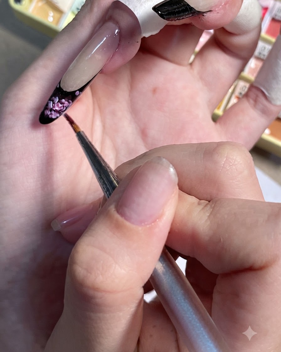 Ein Detailpinsel setzt kleine hellrosa Akzente auf die schwarze French Tip eines lackierten Nagels.