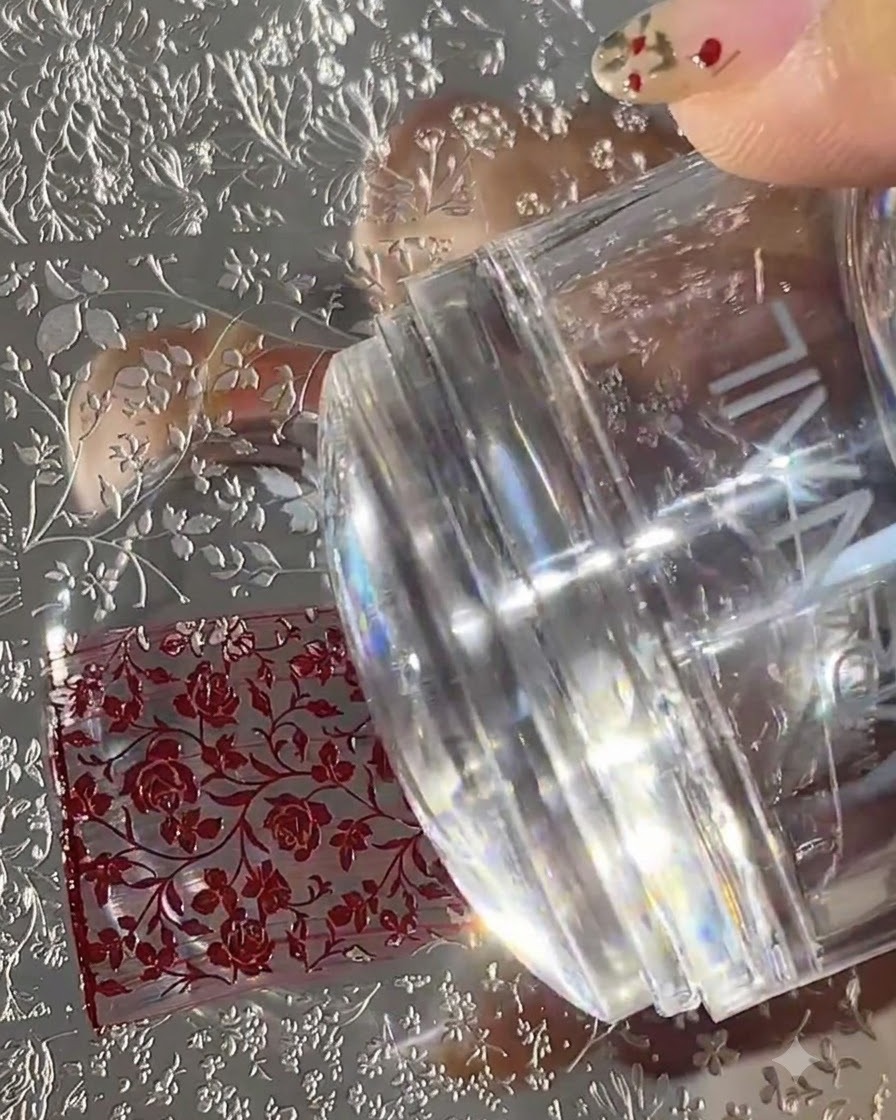 Ein klarer Silikon-Nagelstempel drückt nach unten auf eine gravierte Metall-Stamping-Platte, die mit roten Blumenmustern gefüllt ist.