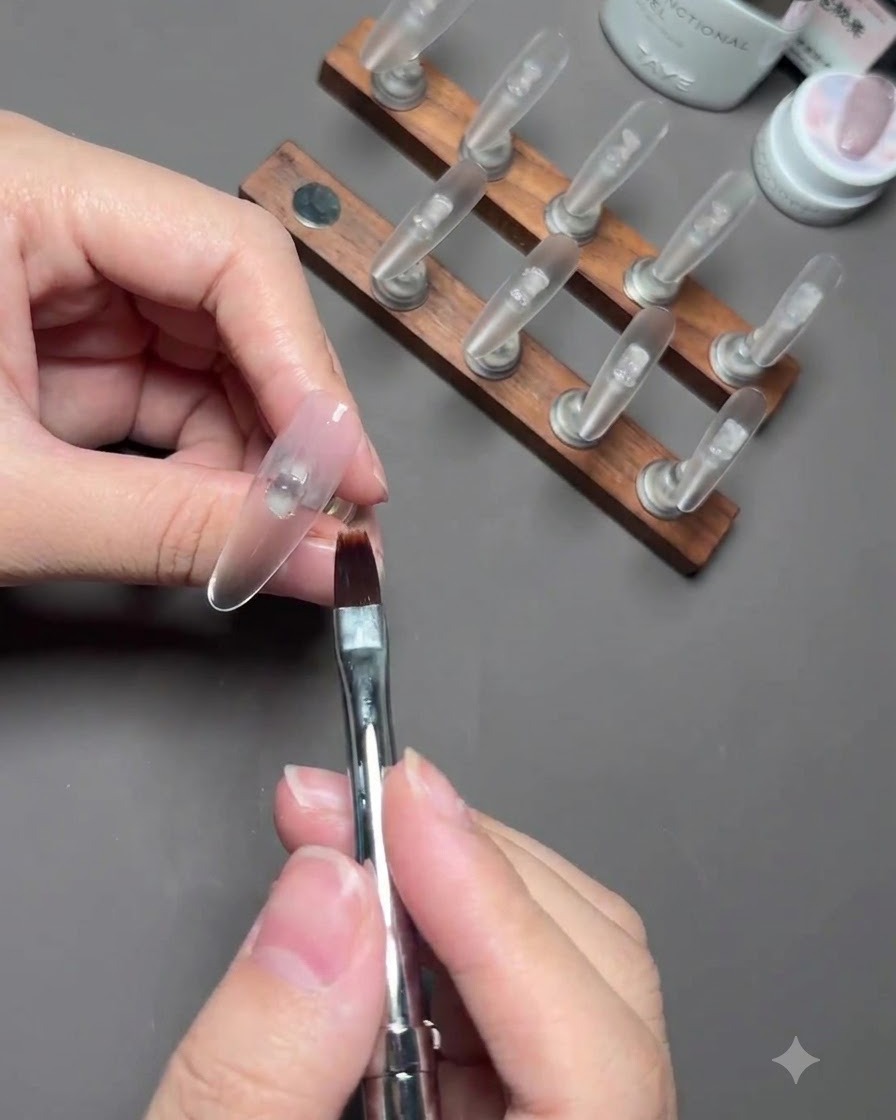 Ein Pinsel verblendet zartes Nude-Farbgel auf einem Daumennagel, der auf einem Ständer befestigt ist.