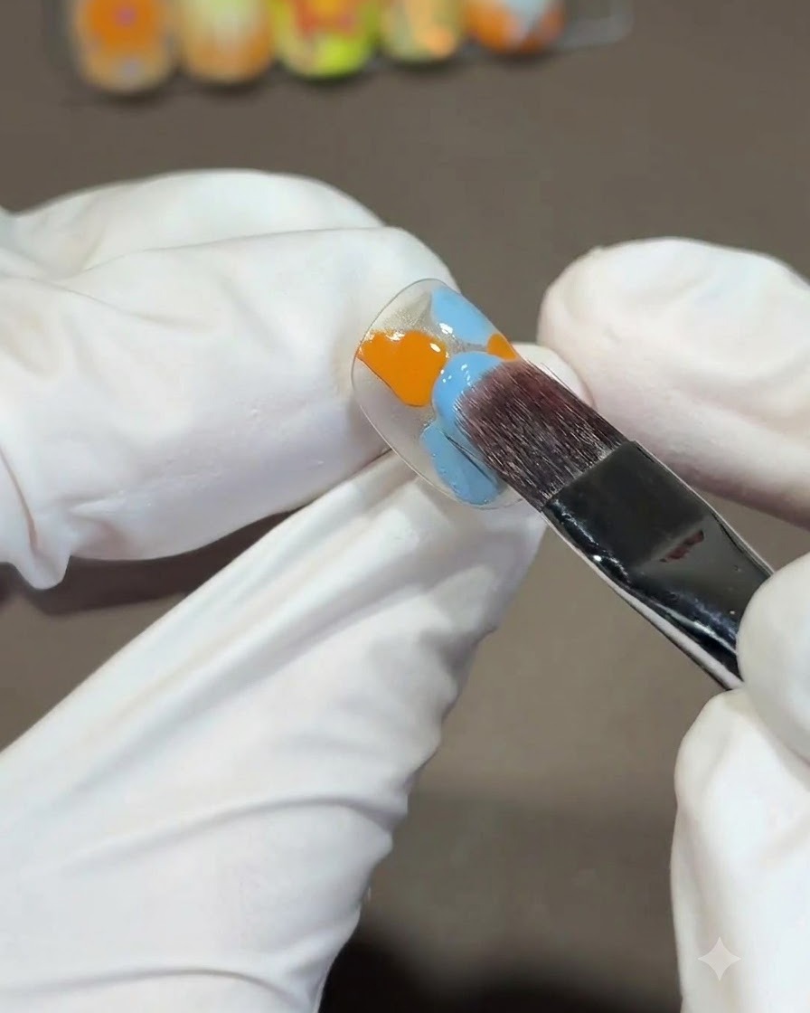 Ein großer, quadratischer Pinsel verblendet orangefarbene und blaue Gel-Polishes auf einer Nagelspitze mit einer S-förmigen Abwärtsbewegung.