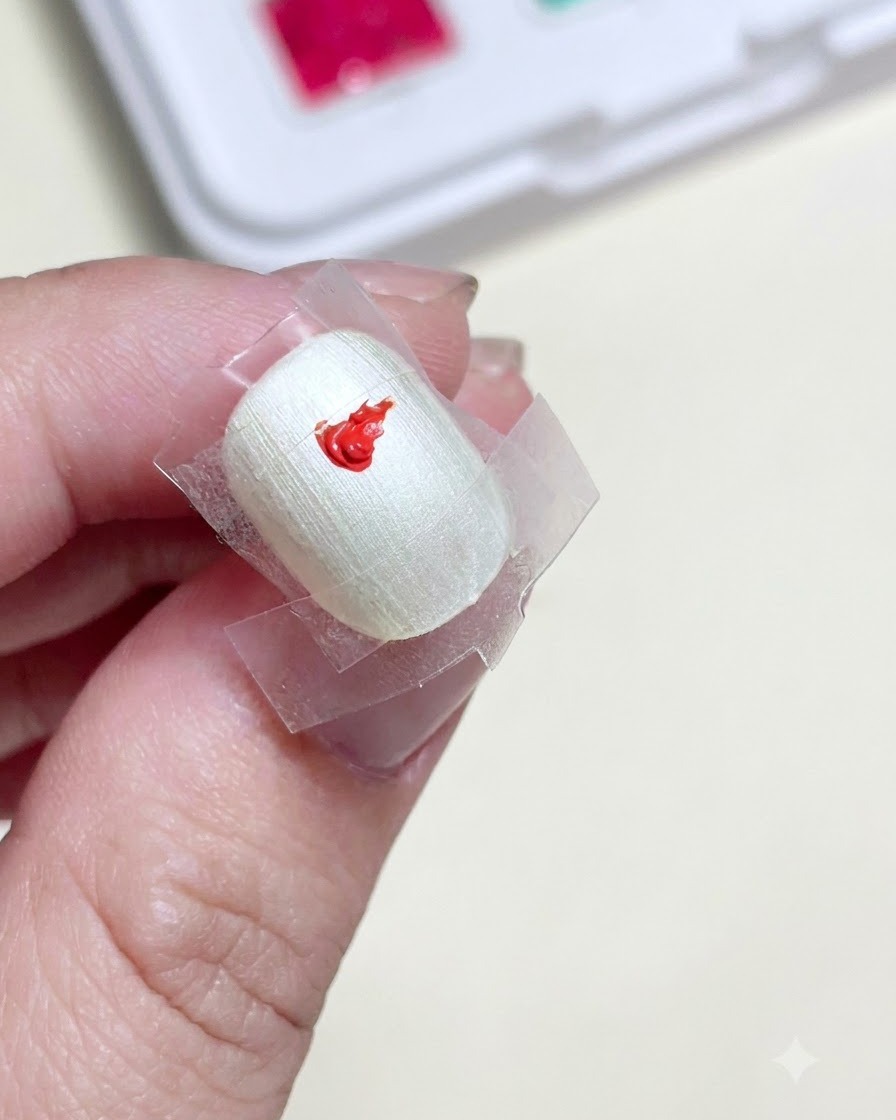 Nahaufnahme eines vorbereiteten Nagels mit einem abgeklebten Quadrat in der Mitte, in dem ein leuchtender Tupfer rotes Farbgel zu sehen ist.