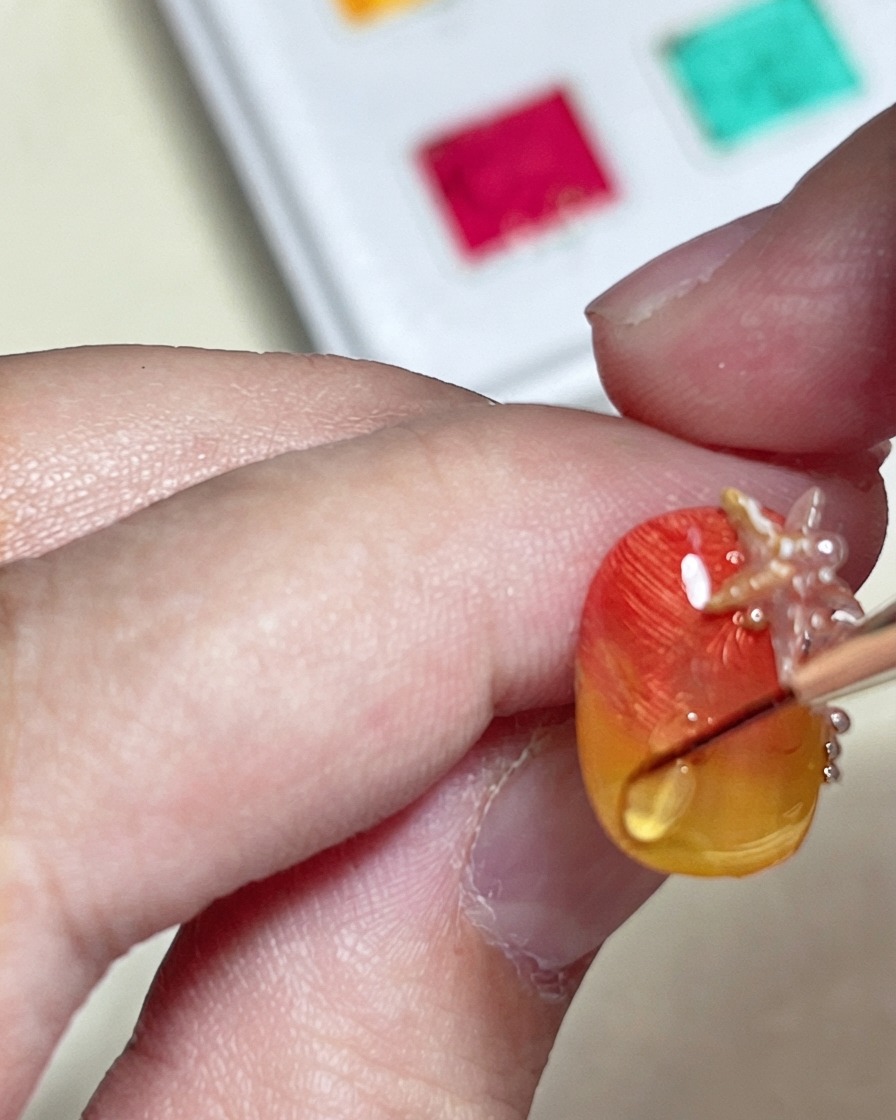 Ein Pinsel zeichnet mit einem Hard Top Coat eine erhabene, glänzende Wasserwelle über einen strukturierten orange-gelben Farbverlaufsnagel.