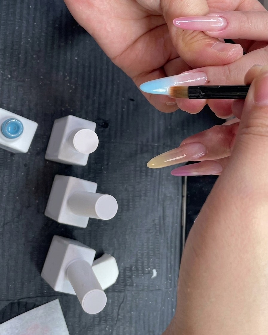 Ein Ombré-Verblenderpinsel wird flach auf einen Fingernagel gehalten, um einen hellblauen Gel-Farbverlauf nach unten zu verblenden.