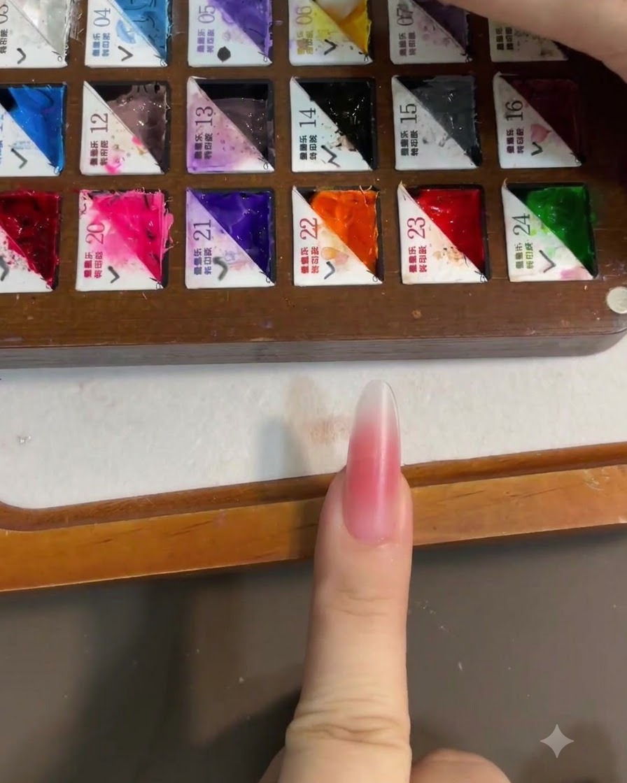 Nahaufnahme eines Pinsels, der hellgelbes Gel-Polish von einer partitionierten Farbpalette aufnimmt, mit einem rosa Blush-Nagel im Vordergrund.