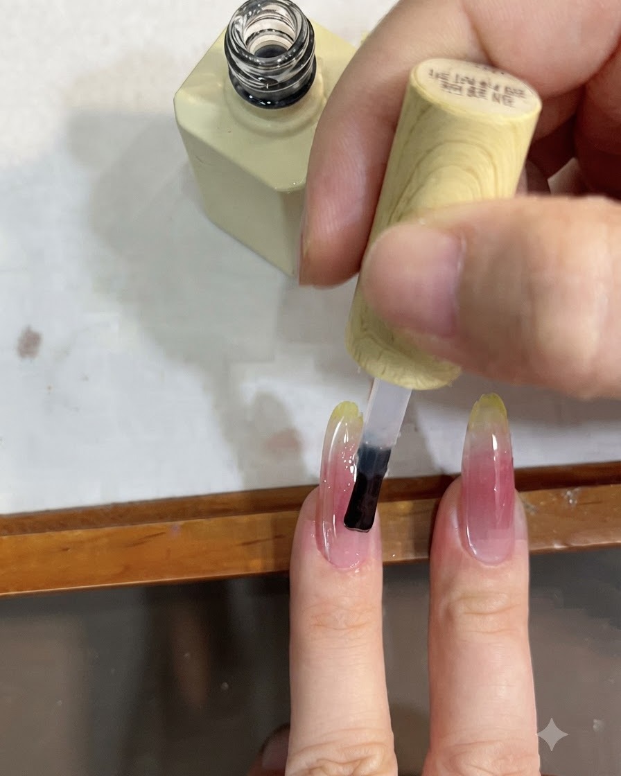 Eine Nahaufnahme eines Pinsels, der einen klaren Top Coat über einen langen Nagel mit rosa und gelbem Farbverlauf aufträgt.