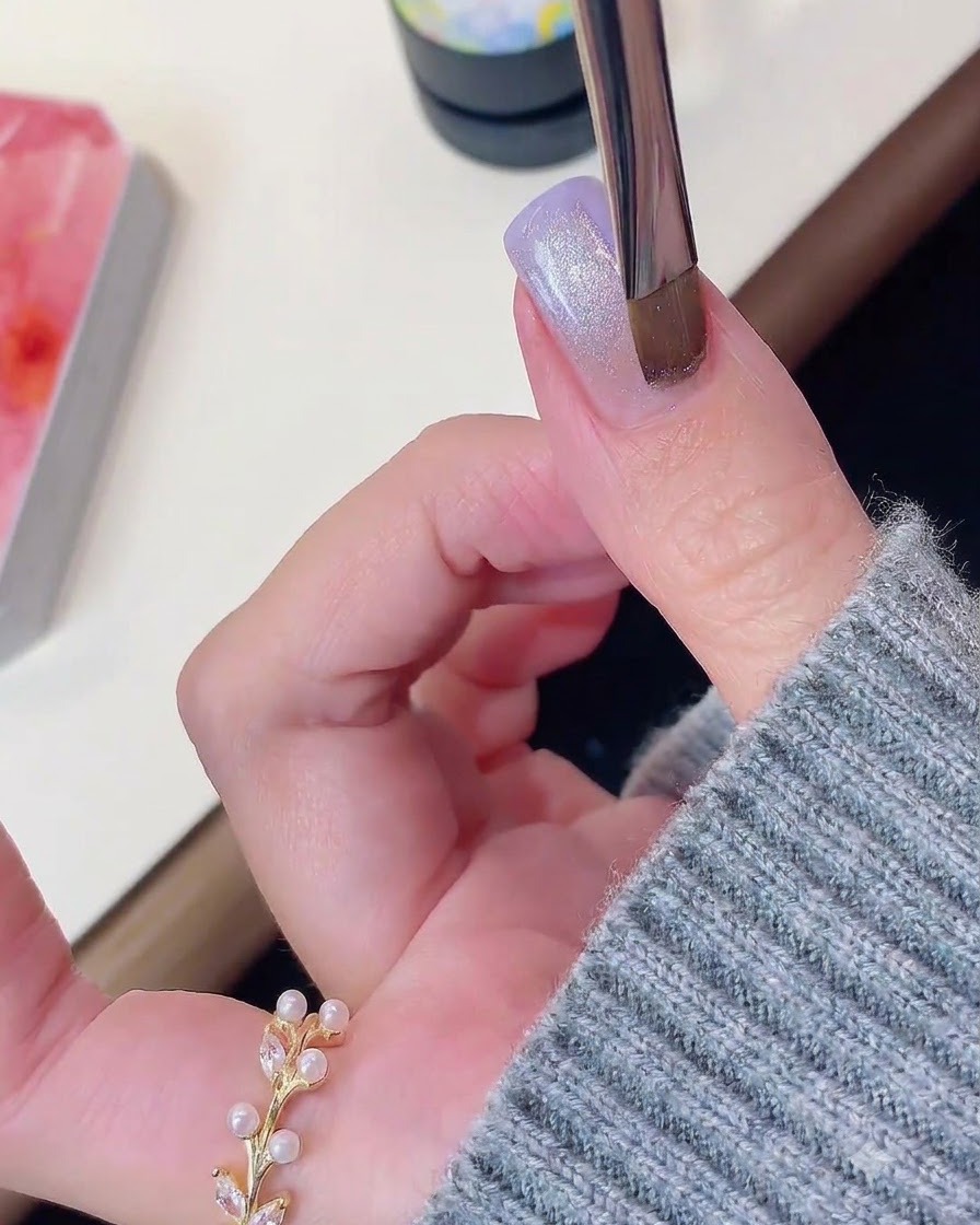Ein Nagelpinsel oder Magnetwerkzeug schwebt über einem lila und schimmernden Cat-Eye-Gel-Nagel.