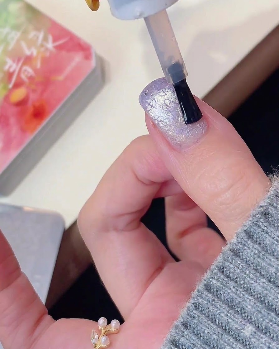 Ein Nagelpinsel trägt Top Coat über ein lila Cat-Eye- und gestempeltes Nageldesign auf.
