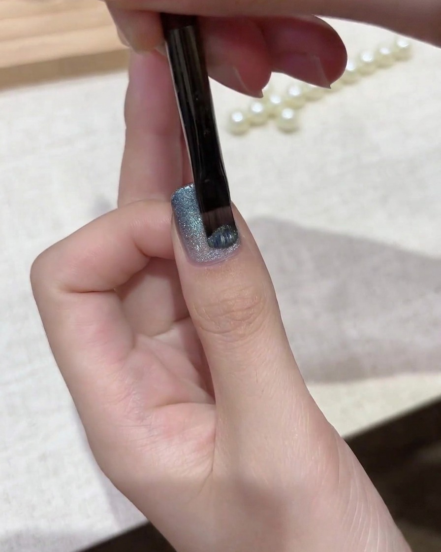 Ein Gelpinsel trägt eine Schicht schimmernden silber-blauen magnetischen Cat-Eye-Lacks über eine Verlaufsgrundierung auf.