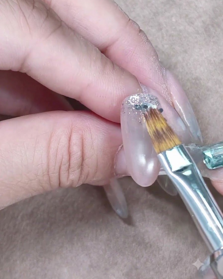 Ein kleiner Nagelpinsel zieht silbernen Glitzer auf einem transparenten Gelnagel sanft nach unten.