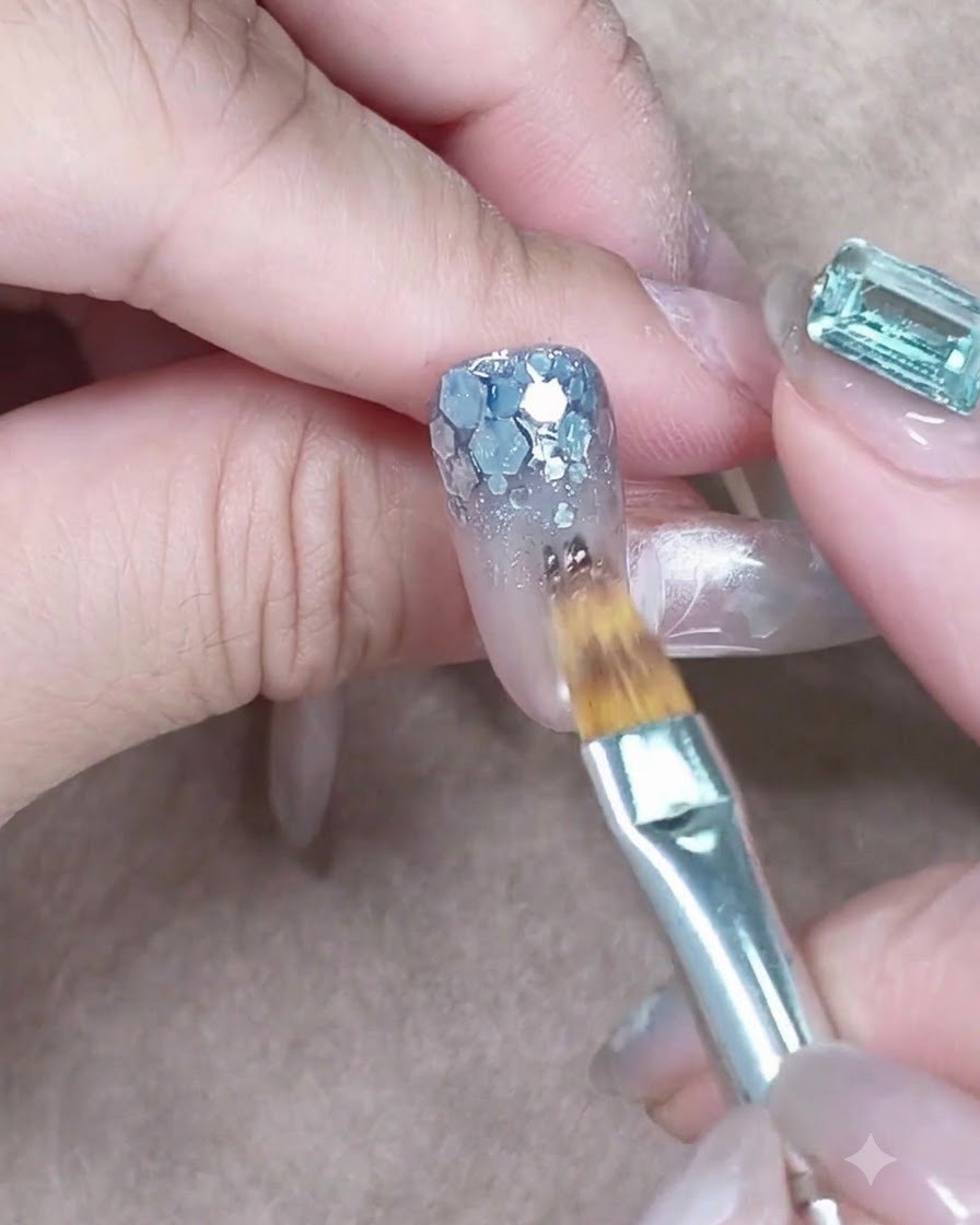 Ein flacher Pinsel tupft transparentes Blau-Gel auf grobe silberne Pailletten am Nagelansatz.