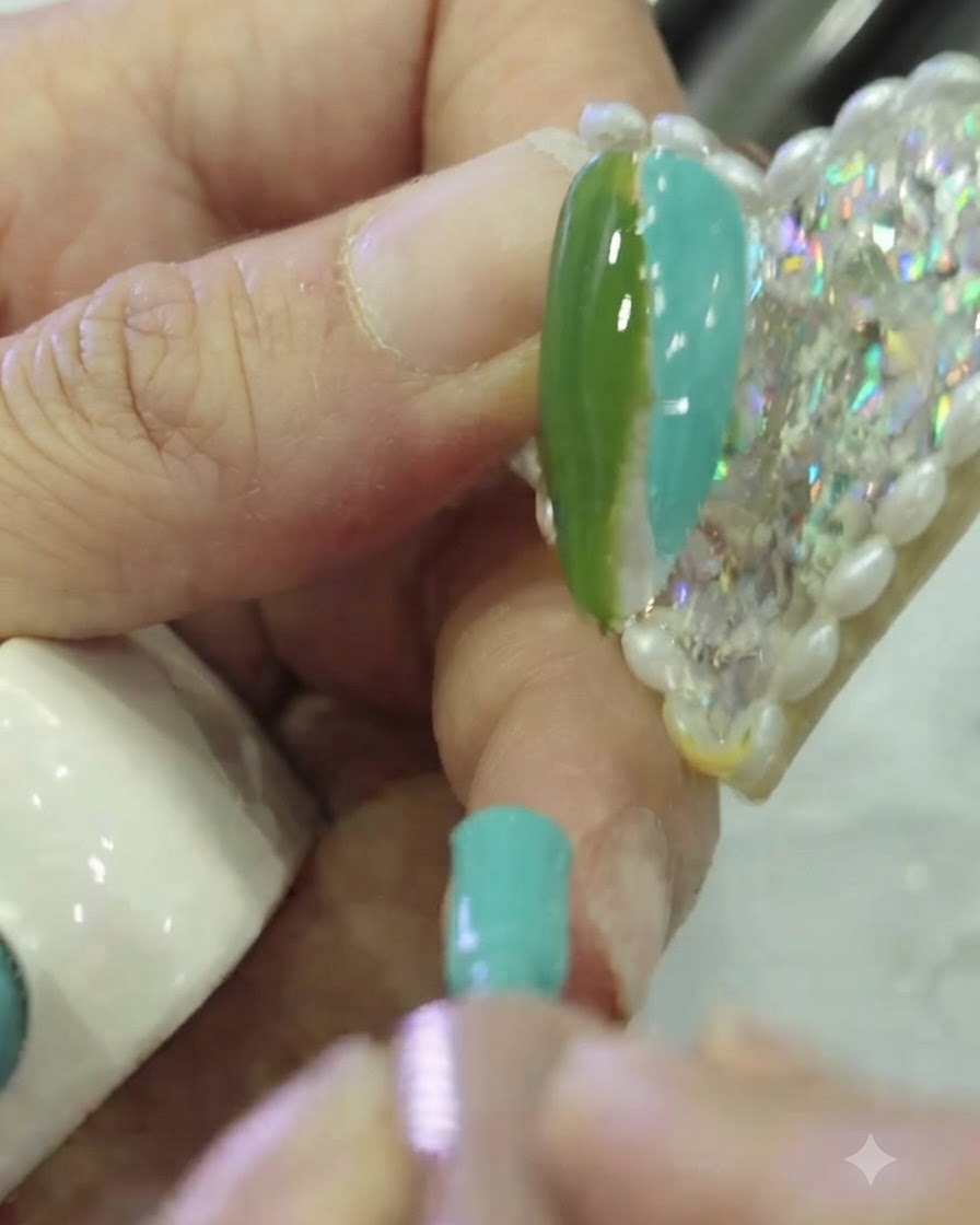 Eine Nahaufnahme eines flachen Nail-Art-Pinsels, der die Grenze zwischen dunkelgrünem und petrolfarbenem Gelack auf einem Nagel abtupft.