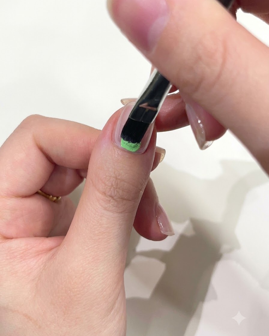 Nahaufnahme einer Hand, die mit einem flachen Gelpinsel mintgrünes Solid Gel auf die untere Hälfte des Daumennagels aufträgt.