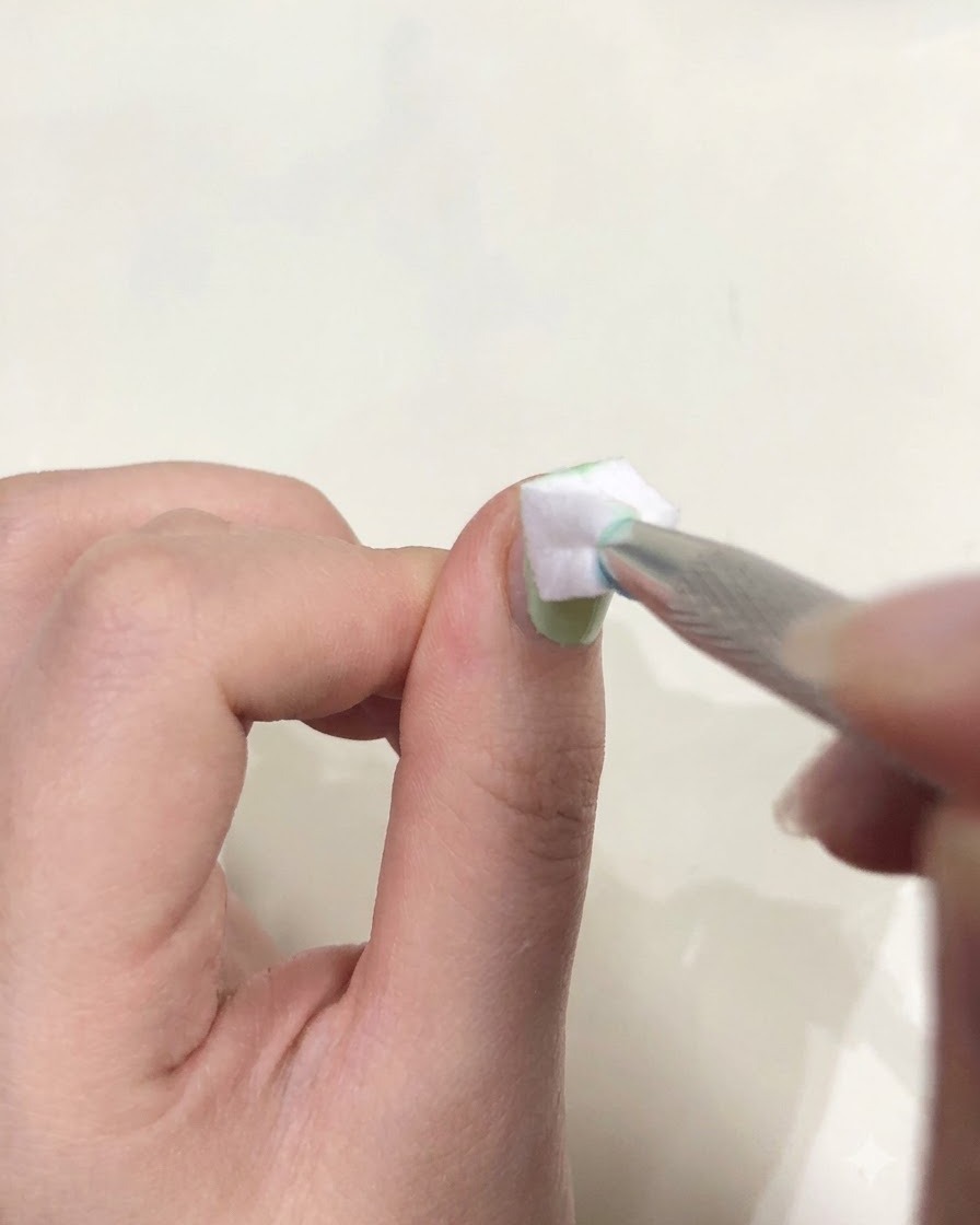 Nahaufnahme einer Hand, die mit einer Pinzette ein kleines weißes Schwämmchen hält und grünes Gel auf einem Fingernagel tupft.