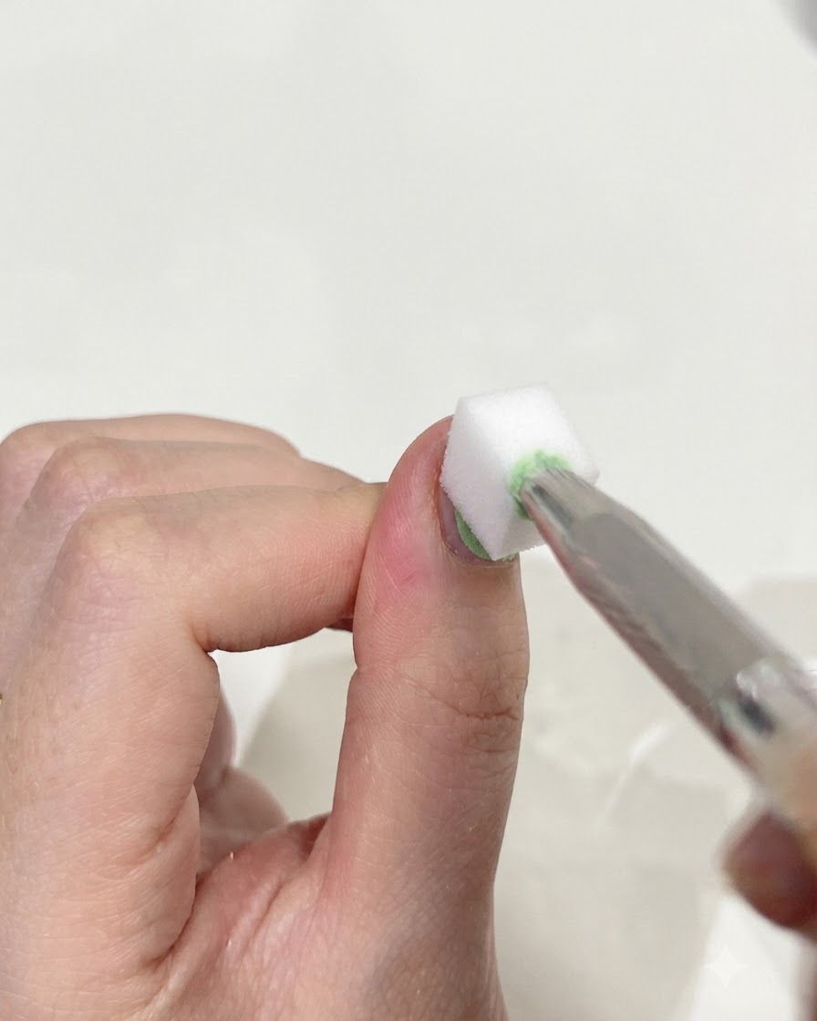 Nahaufnahme einer Pinzette, die ein kleines weißes Make-up-Schwämmchen hält und sanft auf die Oberfläche eines Daumennagels tupft, um grünes und blaues Gel miteinander zu verblenden.