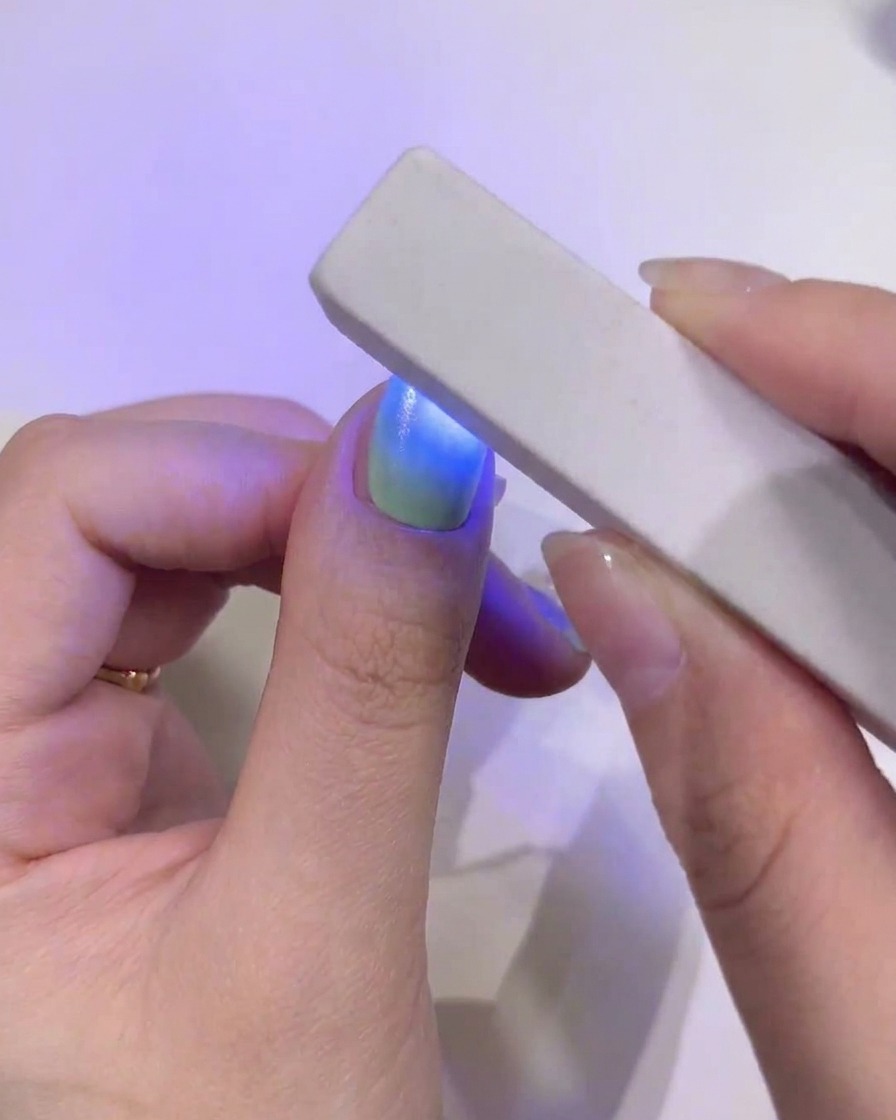 Nahaufnahme eines Daumennagels mit einem nahtlosen grün-blauen Farbverlauf, der unter einer rechteckigen, tragbaren blauen UV/LED-Lampe ausgehärtet wird.