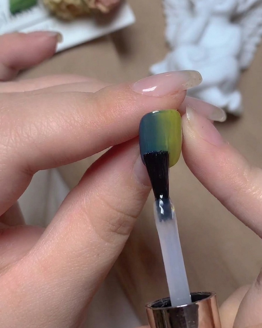 Ein Nagelpinsel trägt einen klaren Top Coat unter warmem Studiolicht auf einen blau-grünen Verlaufsmagel auf.
