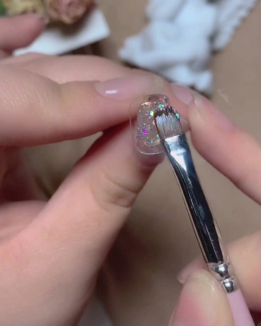 Ein feiner Pinsel trägt glitzerndes, mehrfarbiges Glitzer-Gel mit lebendigen Reflexionen auf eine Nageloberfläche auf.