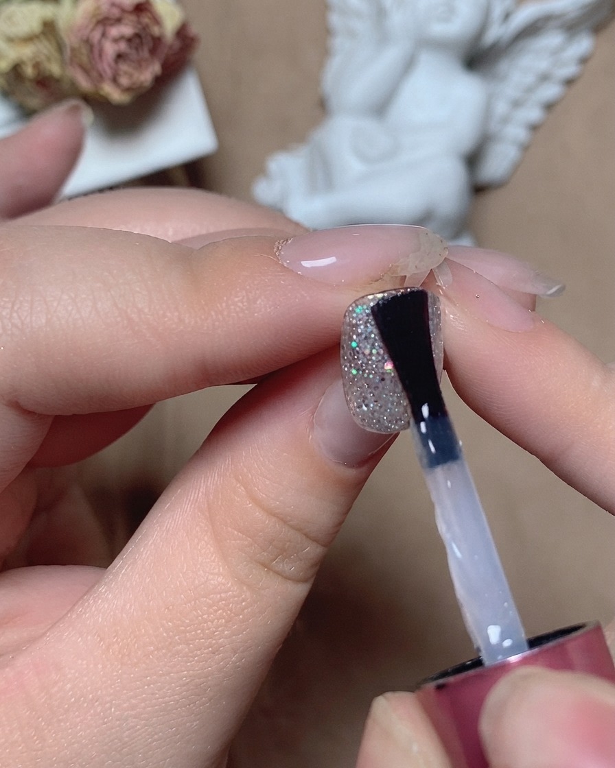 Ein Pinsel trägt unter Studiolicht einen klaren, ausgleichenden Top Coat über einen strukturierten Glitzernagel auf.
