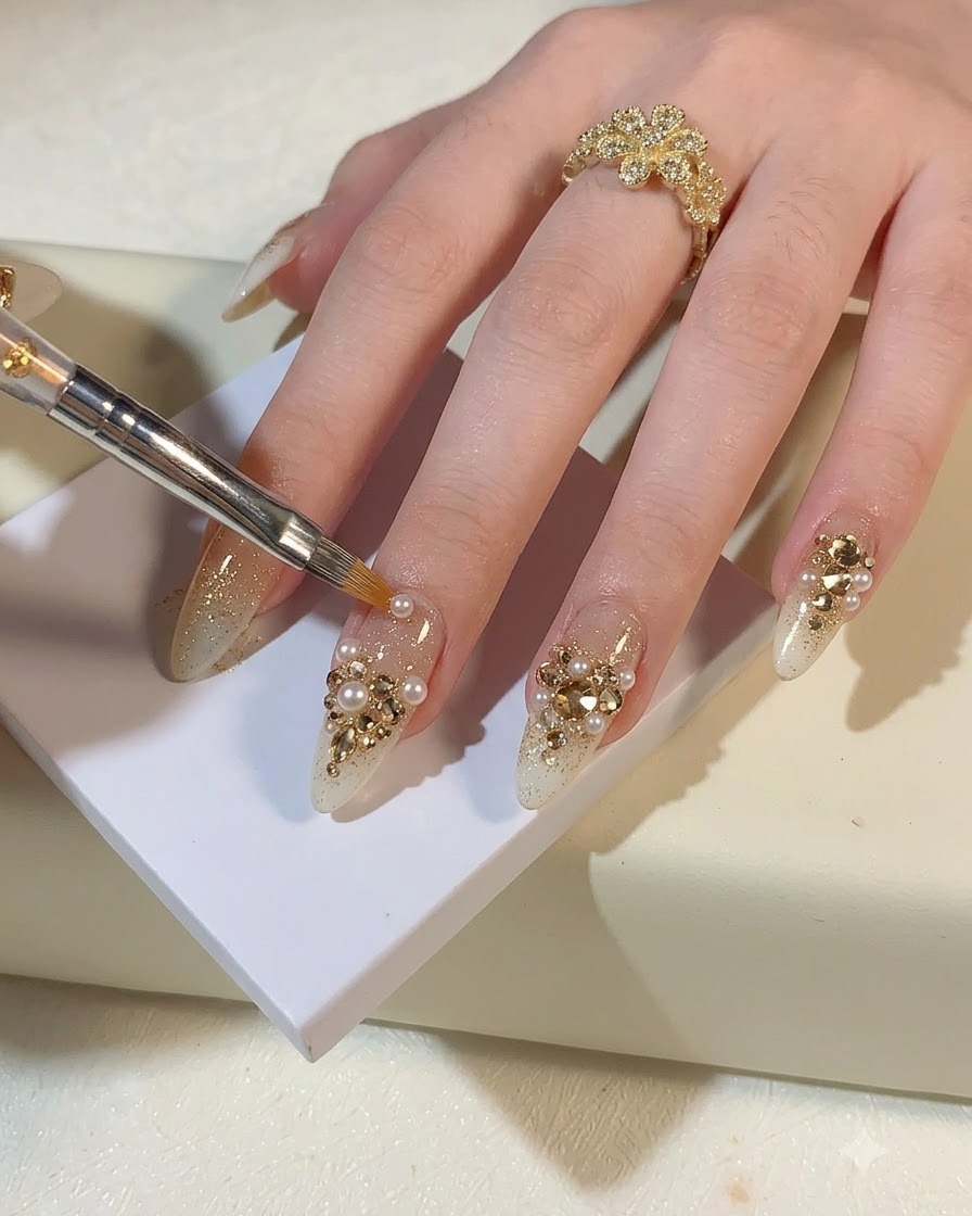 Ein Detailpinsel oder Werkzeug platziert kleine Perlen und champagnergoldene Strasssteine auf einem Glitzernagel.
