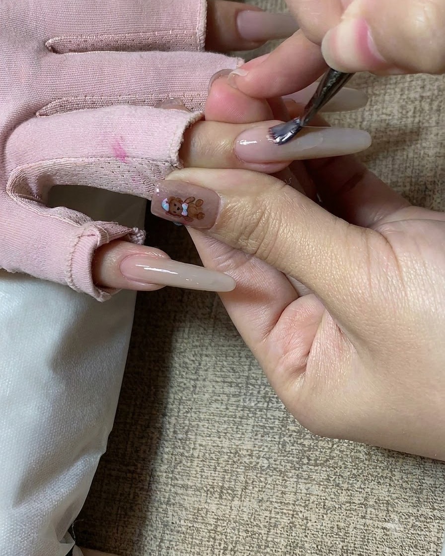 Ein Nagelpinsel trägt milchteefarbenes Gel auf einen langen, modellierten Fingernagel auf.cite: 1