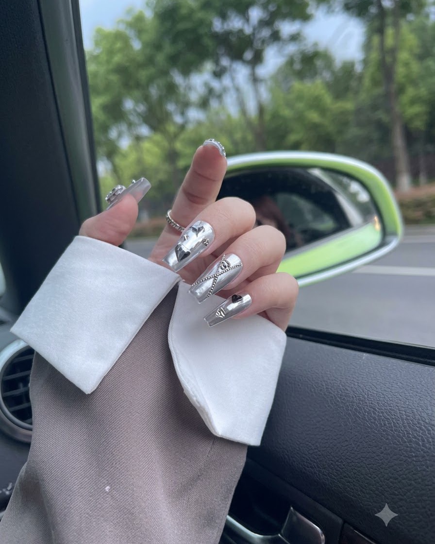 Finger bewegen sich sanft im Tageslicht und zeigen silberne Metallic-Press-On-Nägel mit baumelnden Ketten.