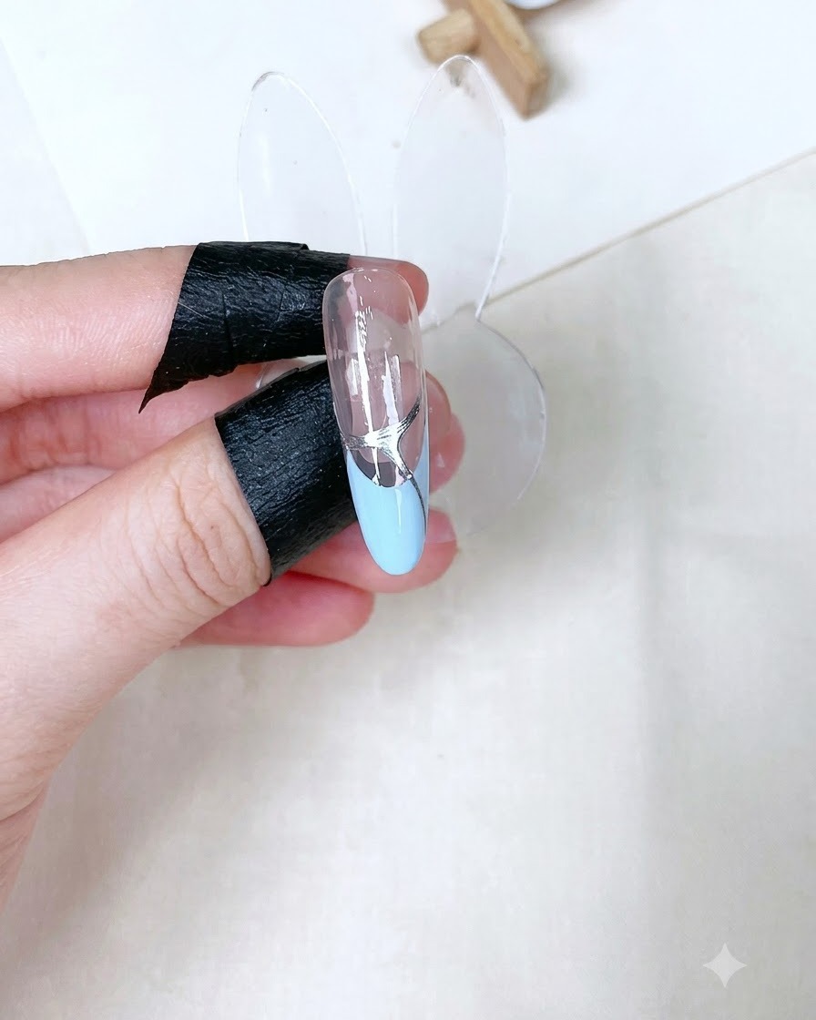 Ein einzelner modellierter Nagelstand zeigt einen ausgehärteten, glänzenden Top Coat mit silbernen Details über einer babyblauen French-Spitze.