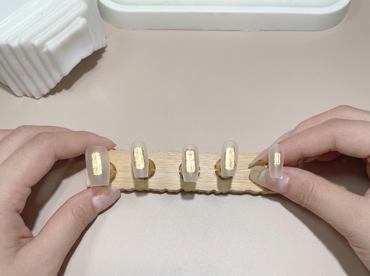 Eine Nahaufnahme eines Pinsels, der Base Gel auf mehrere auf einem Holzständer befestigte Nagelspitzen aufträgt.